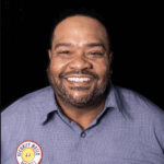 Headshot of JJ Campbell in a blue button-down security water shirt, smiling in front of a dark background
